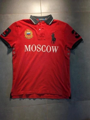 Röd Polo Ralph Lauren Moscow piké - Röd pikétröja från Polo Ralph Lauren med svart krage och detaljer. Stort tryck 'MOSCOW' framtill, broderad logga och emblem, samt siffran 3 på ärm och rygg. Klassisk och sportig stil med korta ärmar.