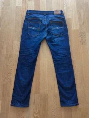 Mörkblå jeans från Nudie Jeans - Säljer ett par mörkblå jeans från Nudie Jeans med klassisk femficksdesign och raka ben. Jeansen har kontrastsömmar och subtila slitningar för en snygg look. Perfekta för dig som gillar stilrena och tidlösa jeans i denim. 32/32