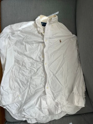 Vit skjorta från Polo Ralph Lauren - Klassisk vit skjorta från Polo Ralph Lauren med broderad logga på bröstet. Skjortan har knappar hela vägen fram, lång ärm och button-down krage. Skjortan har en fläck i nacken som syns på tredje bilden.