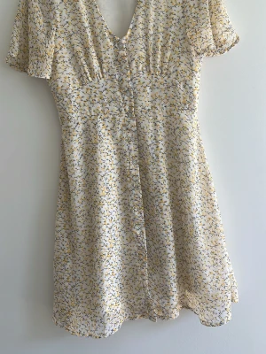 Blommig kortklänning - Söt kortklänning från Vero moda med blommigt mönster i gult och blått på vit botten. Klänningen har v-ringning, knappar framtill och korta ärmar. Perfekt för sommarens dagar, lätt och luftig. Går ungefär lite över knäna 