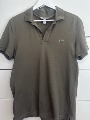 Olivgrön piké från Lacoste - Klassisk olivgrön pikétröja från Lacoste med korta ärmar och den ikoniska krokodilloggan på bröstet. Tröjan har krage och knappar vid halsen. Tillverkad i bomull för en skön och luftig känsla. Perfekt skick, säljer då den är för liten.