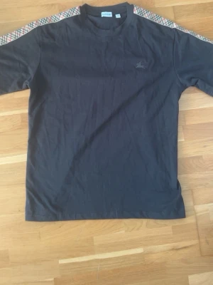 Svart Burberry t-shirt med rutiga detaljer - Snygg svart t-shirt från Burberry med klassiska rutiga paneler längs axlarna och ärmarna. T-shirten har rund halsringning och ett diskret broderat Burberry-logotyp på bröstet. Tillverkad i mjuk bomull för skön känsla.