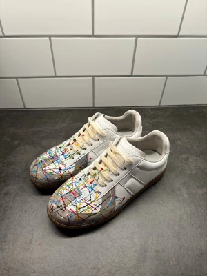 Maison margiela skor - Unika vita sneakers med färgstänk ifrån maison margiela. Skorna har klassisk låg silhuett, vita snören och sula i gummi. Materialet är läder och skorna är iprincip helt nya och använts fåtal gånger. Skorna kommer tyvärr inte med box eftersom jag slängde den när jag fick skorna. Storleken är 40 men dem är ganska breda. Varan skickas inom 24t om möjlighet😁. Hör av er vid frågor!