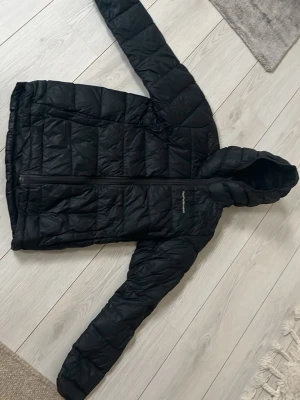 Svart dunjacka Peak Performance - Snygg svart dunjacka från Peak Performance med huva och dragkedja framtill. Jackan har quiltad design och diskret logga på bröstet. Perfekt för kalla dagar och enkel att matcha med olika outfits.
