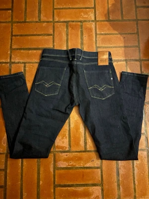 Mörkblå jeans från Replay - Säljer ett par mörkblå jeans från Replay med raka ben och klassisk femficksmodell. Jeansen har kontrastsömmar och detaljerade bakfickor med snygg brodyr. Tillverkade i kraftig denim som ger en stilren look.