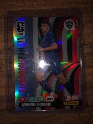 Abdukodir Khusanov FIFA 2026 kort - Samlarbild från Panini Adrenalyn FIFA World Cup 2026 med spelaren Abdukodir Khusanov. Kortet har holografisk yta med färgglada detaljer och texten 'FAN FAVOURITE'. Kortet är inplastat och har statistik och spelarinfo på framsidan.