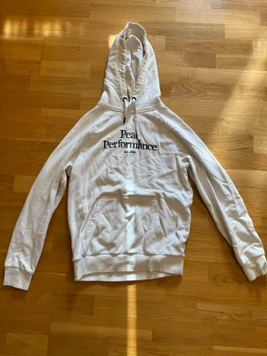 Vit hoodie från Peak Performance - Snygg vit hoodie från Peak Performance med svart broderad logga och text 'Est. 1986' på bröstet. Klassisk modell med huva, dragsko och känguruficka framtill. 
