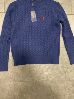 Polo Ralph Lauren Tröja - Snygg blå kabelstickad tröja från Polo Ralph Lauren med rund halsringning och klassisk röd logga broderad på bröstet. Tröjan har långa ärmar och ribbade muddar vid ärmslut och nederkant. Perfekt för en stilren och avslappnad look. Den har använts ett par gånger så taggarna är inte kvar, men dem är i mycket bra skick.