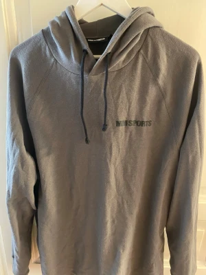 Grå hoodie från MM Sports - Snygg grå hoodie från MM Sports med svart logga på bröstet. Hoodien har dragsko i huvan och är tillverkad i ett mjukt, strukturerat material. Perfekt för en avslappnad och sportig stil.