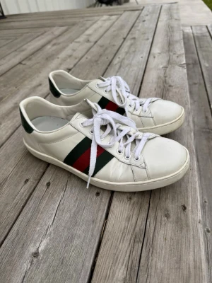 Gucci vita sneakers med gröna detaljer - Snygga vita sneakers från Gucci med klassiska gröna och röda ränder på sidan. Skorna har gröna detaljer i läder baktill och vita snören. Tillverkade i läder med låg profil och stilren design som passar till många outfits.storlek 40 mer som 41-42