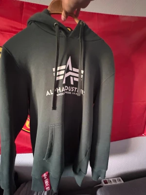 Mörkgrön hoodie från Alpha Industries - Snygg mörkgrön hoodie från Alpha Industries med vit logga och text på bröstet. Klassisk känguruficka framtill, dragsko i huvan och ribbade muddar. Perfekt för en avslappnad streetwear-look.