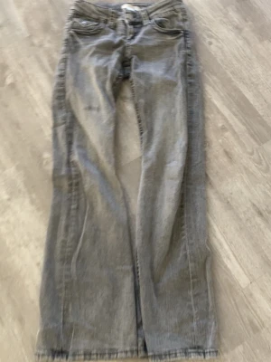 Grå bootcut jeans med fickdetaljer - Säljer ett par grå bootcut jeans med klassisk femficksdesign och snygga ficklock med knappar bak. Jeansen har normal midja och raka ben. Perfekta för en avslappnad och trendig look. Materialet är denim i bomull.