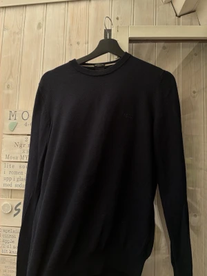 Svart stickad tröja från BOSS - Snygg svart stickad tröja från BOSS med diskret logga på bröstet. Tröjan har rund halsringning och långa ärmar. Perfekt basplagg i garderoben och passar till det mesta. Materialet känns mjukt och följsamt. Finns inga dumma frågor, priset  hugget i sten