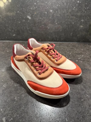 Loro Piana sneakers - Snygga sneakers med beige textil, orange och vinröda mockadetaljer samt matchande vinröda skosnören. Skorna har en chunky vit sula och en modern, sportig look. Perfekta för dig som vill sticka ut med färg och stil. Ingen synlig logga eller märke.