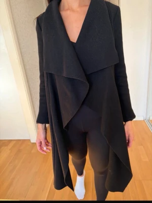 Svart draperad kappa med öppen front - Stilren svart kappa med lång, draperad och öppen front. Kappan har långa ärmar och ett mjukt, följsamt tyg som ger ett snyggt fall. Perfekt för lager på lager och en modern look. Passar till både jeans och klänning.passar S-m