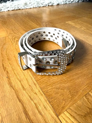 Vitt bälte med nitar och strass - Snyggt vitt bälte med massor av silvriga nitar och glittrande strassdetaljer på spännet.  Bältet har hål längs hela längden och ett stort, glittrigt spänne i silver. Aldrig använt 😊