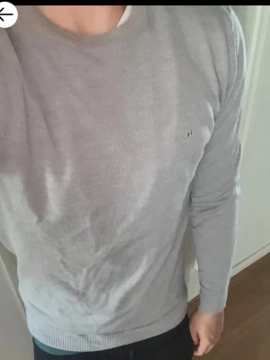 Ljusgrå tröja från J.Lindeberg - Snygg ljusgrå tröja från J.Lindeberg i tunn, mjuk bomull. Klassisk rund halsringning och diskret logga på bröstet. Perfekt till jeans eller chinos för en clean och stilren look.