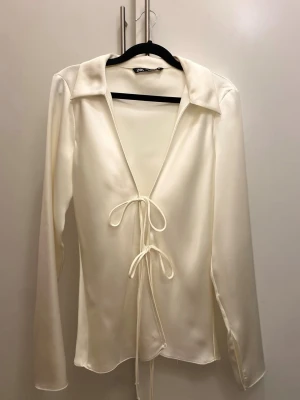 Viral krämvit satinblus från Zara - Stilren krämvit blus från Zara i glansigt satinmaterial. Blusen har djup V-ringning, långa ärmar och knytdetaljer framtill. Perfekt för en uppklädd elegant look med bred krage och mjukt fallande tyg, men funkar också till en mer vardags/basic outfit med t.ex jeans. Storlek M
