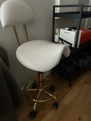 Vit frisör/nagel/frans stol med GULDDETALJ  - Säljer en vit snurrstol med ryggstöd och guldfärgade metallben. Stolen har hjul för enkel förflyttning och sitsen är klädd i vitt konstläder. Perfekt till hobby verksamhet eller annat. Pris kan diskuteras då jag behöver få den såld pga flytt 6/6. FINNS I BARKARBY 