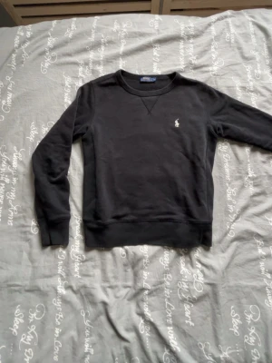 Svart sweatshirt från Polo Ralph Lauren - Klassisk svart sweatshirt från Polo Ralph Lauren med rund halsringning och broderad vit logga på bröstet. Tröjan har ribbade muddar vid ärmslut och nederkant samt mjuk insida för extra komfort. Perfekt för en avslappnad och stilren look.