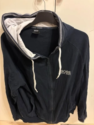 Marinblå hoodie från Hugo Boss - Snygg marinblå hoodie från Hugo Boss med dragkedja framtill och vit logga på bröstet. Huvan har vit insida och vita snören, vilket ger en schysst kontrast. Tillverkad i mjukt bomullsmaterial och har två fickor framtill. Perfekt för en avslappnad och stilren look.