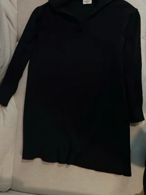 Svart långärmad stickad klänning Chiquelle - En svart långärmad stickad klänning från Chiquelle med krage och enkel, rak passform. Klänningen är kort och har en stilren look som passar till många olika tillfällen. Perfekt för dig som gillar minimalistisk stil.