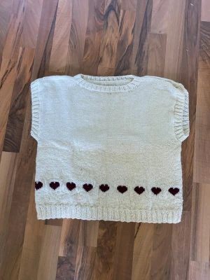 Stickad väst med hjärtmönster - Söt stickad väst i ljusbeige med mörkröda hjärtan längs nederkanten. Västen har ribbstickade muddar vid hals, ärm och nederkant. Perfekt att slänga över en skjorta eller t-shirt för en gullig look. 100% ull