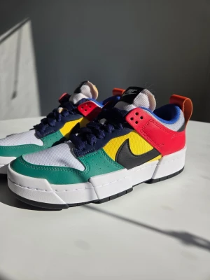 Nike Dunk Low Disrupt multi-color - Färgglada Nike sneakers med vit mesh, gröna och röda läderpaneler, gul textil, blå detaljer och svart Swoosh. Skorna har chunky vit sula, marinblå snören och orange hälflik. Perfekt för dig som gillar streetstyle och unika färgkombinationer.