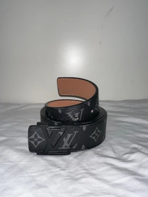  svart bälte med L&V-Spänne - Snyggt svart skärp med klassiskt monogrammönster och -spänne i svart metall. Bältet är gjort i skinn med beige insida och har en bred design som sticker ut.