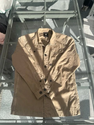 G-Star RAW Overshirt Beige L – Utility / Workwear - Säljer en snygg G-Star RAW overshirt i beige färg, storlek L. Modellen har en stilren utility- och workwear-inspirerad design med flera fickor och en rak passform som gör den perfekt att bära över t-shirt, skjorta eller hoodie. Ett mångsidigt plagg som fungerar lika bra till jeans som cargo pants och passar året runt.  Den neutrala beige färgen och den tidlösa designen gör detta till ett självklart val för dig som gillar streetwear, casual och modern herrstil. Plagget är i Mycket bra skick.