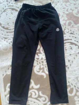 Svarta mjukisbyxor från Moncler - Säljer ett par svarta mjukisbyxor från Moncler med elastisk midja och diskret logga på vänster sida. Byxorna har raka ben och är tillverkade i mjukt material som känns skönt mot huden. Perfekta för chill dagar eller när du vill ha en clean look.