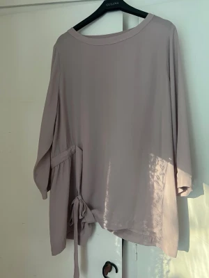 Blus från Åhléns - Säljer en ljuslila oversized blus från Åhléns med rund halsringning och långa ärmar. Blusen har ett dekorativt knytband som man knyter baktill och är tillverkad i ett mjukt, tunt tyg som faller snyggt. Perfekt för en avslappnad men stilren look.