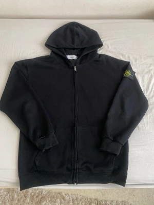 Svart hoodie från Stone Island - Säljer en svart hoodie från Stone Island med dragkedja och huva. Klassiskt logopatch på vänster ärm. Hoodien har ribbade muddar och är tillverkad i mjukt material, perfekt för en avslappnad och cool look. Passform mellan M och L 