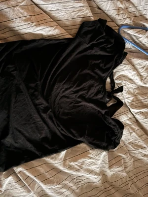 Svart topp med rosett i satinband - Snygg svart topp med korta ärmar och en dekorativ rosett i satinband bak i nacken. Toppen har en enkel, klassisk design och är tillverkad i ett mjukt material som känns skönt mot huden. Perfekt för dig som gillar stilrena plagg med en liten twist.