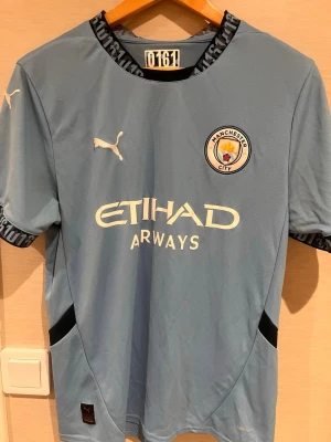 Manchester City matchtröja Puma - Ljusblå Manchester City matchtröja från Puma med klubbmärke och Etihad Airways-tryck på bröstet. Tröjan har svarta och blå detaljer vid krage och ärmslut samt korta ärmar. Perfekt för fotbollsträning eller att bära på matchdag.
