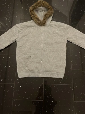 Grå hoodie med fuskpäls i huvan - Snygg grå zip-hoodie med mjuk fuskpäls i huvan. Klassisk modell med dragkedja framtill, två fickor och ribbade muddar. Perfekt för en chill och avslappnad stil. Huvan har en fluffig kant som ger extra cozy känsla.