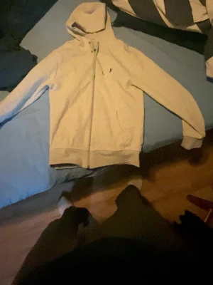 Grå hoodie från Polo Ralph Lauren - Snygg grå hoodie från Polo Ralph Lauren med dragkedja och klassisk huva. Liten broderad logga på bröstet och ribbade muddar vid ärmslut och nederkant. Tillverkad i mjukt bomullsmaterial, perfekt för en avslappnad stil.