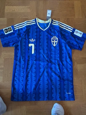 Blå Sverige retro fotbollströja Adidas - Retroinspirerad blå fotbollströja från Adidas med Sveriges landslagsemblem, nummer 7 och vita detaljer. Tröjan har klassiska Adidas-ränder på axlarna, V-ringning med vit kant och mönstrad struktur i tyget. Perfekt för fotbollsfans och samlare.