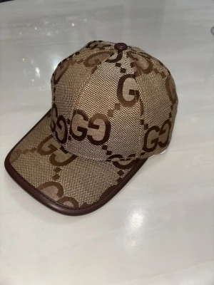  Gucci jumbo  - Snygg beige keps från Gucci med klassiskt brunt GG-monogram över hela kepsen. Kepsen har böjd skärm och bruna detaljer längs kanten. Tillverkad i canvasmaterial med exklusiv känsla och modern streetwear-look.