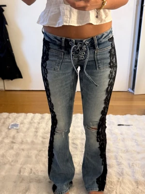 Blå jeans med svart spetsdetalj - Unika blå jeans med snörning framtill och svarta spetsdetaljer längs sidorna jag sytt på. Modellen är bootcut med slitningar på ena benet och klassiska fickor bak. 