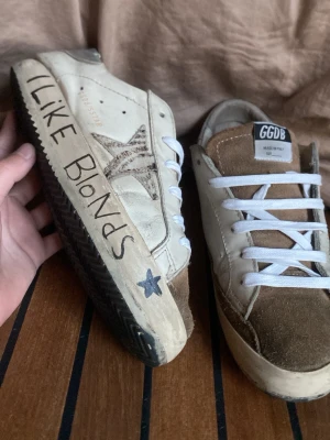 GGDB sneakers med text och stjärna - Unika GGDB sneakers i beige och vitt med handskrivna texter på sulan, stjärndetalj på sidan och kontrasterande bruna partier i mocka. Klassisk låg modell med vita skosnören och GGDB-logga på plösen. Perfekta för dig som gillar personlig stil.
