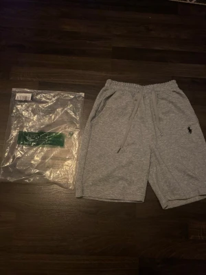 Grå sweatshorts från Polo Ralph Lauren - Grå mjuka sweatshorts från Polo Ralph Lauren med elastisk midja och snörning. Klassisk svart logga broderad på benet och två sidofickor. Perfekta för chill dagar eller träning. Helt nya | Perfekt nu till sommaren🤩