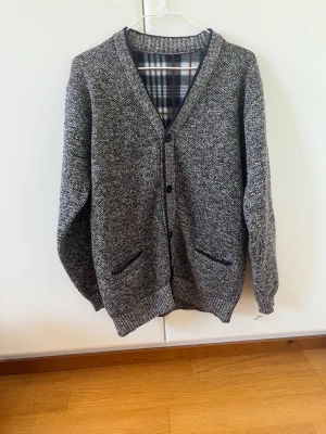 Gråmelerad kofta med knappar - Kofta i gråmelerat stickat material med svarta knappar och två fickor framtill. Insidan har ett rutigt foder i grått, vitt och brunt. Klassisk v-ringad modell med ribbade muddar och nederkant. Perfekt för lager-på-lager och chill stil.