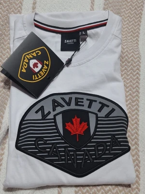 Vit barn t-shirt från Zavetti Canada - Vit barn t-shirt från Zavetti Canada med stort broderat emblem på bröstet i svart och grått, samt röd lönnlöv. Rund halsringning med snygg detaljrand. Perfekt för dig som vill ha en cool och stilren look. Barn st L