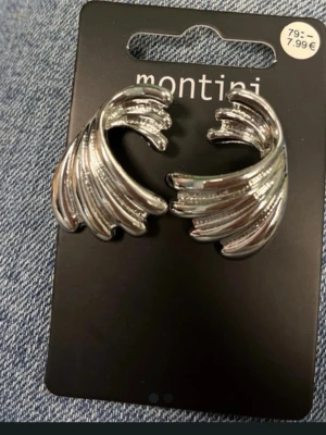Silvriga örhängen från Montini - Snygga silvriga örhängen från Montini med vågformad design. De har en blank finish och är tillverkade i metall. Perfekta för att ge din look en cool och modern touch.