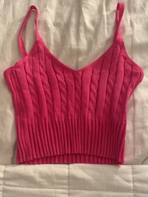 Rosa ribbad stickad linnetopp - Snygg rosa stickad linnetopp med smala axelband och kabelstickat mönster. Perfekt till en skön sommar dag med jeans eller en kjol som passar för en trendig och avslappnad look.