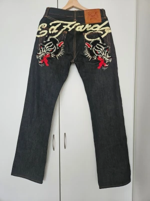 Ed Hardy jeans med drakbrodyr - Säljer ett par Ed Hardy Y2K-jeans från 2009 i mörkblått. Äkta 2000s‑stil med snygg och tydlig brodering. •Mycket fint begagnat skick •Broderingen är hel och klar Mått: Midja: 76 cm Innerben: 87 cm Ytterben: 114 cm Benöppning: 24 cm