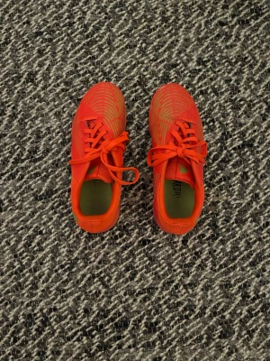 Röda Adidas fotbollsskor inomhus - Säljer ett par röda Adidas fotbollsskor för inomhusbruk. Skorna har gröna detaljer på sidan och snörning framtill. Ovandelen är i syntetmaterial med ventilerande meshpaneler och en låg profil för bra bollkontroll. Perfekta för futsal eller inomhusträning.