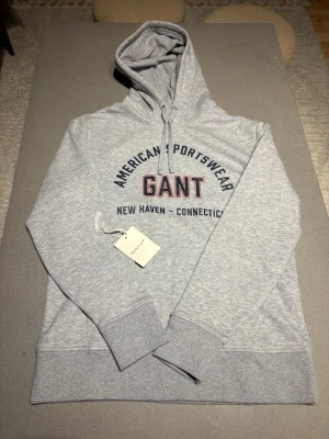 Grå hoodie från GANT med tryck - Snygg ljusgrå hoodie från GANT med huva och dragsko. Tröjan är oanvänd och kostar 1300 kronor ny. Stor logga och texten 'American Sportswear' samt 'New Haven - Connecticut' framtill. Tillverkad i mjuk bomullsblandning.
