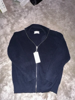 Maison Margiela Zip Tröja - Säljer en mörkblå stickad Maison Margiela tröja med hög krage och dragkedja framtill. Tröjan har ett klassiskt ribbstickat mönster och vita sömdetaljer på ryggen. Perfekt för att hålla sig varm under kyliga dagar.